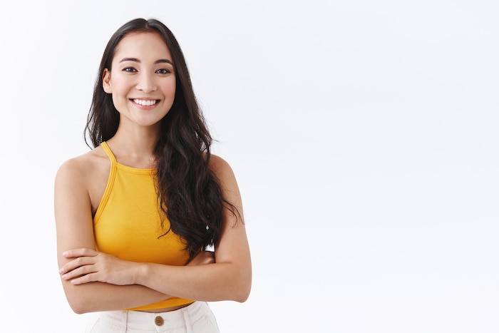 Confident, attractive young outgoing asian woman in yellow top, smiling friendly and happy as cross hands chest, posing over white background self-assured, sassy pose, look determined