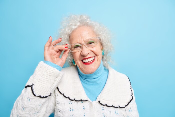 Portrait of good looking cheerful grey haired grandmother smiles toothily keeps hand on rim of spectacles has well cared complexion wrinkled sin dressed in white jumper