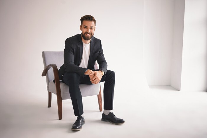 People, lifestyle, business, style, fashion and men\'s wear concept. positive successful young ceo sitting in armchair, smiling, dressed in elegant shoes, trousers, jacket and white t-shirt