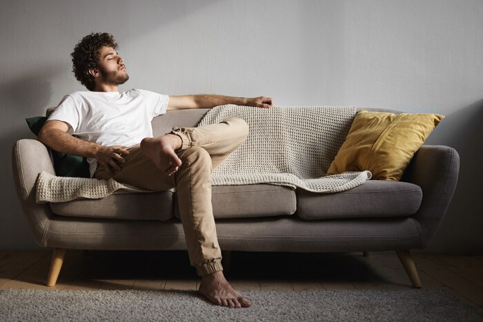 Picture of stylish handsome young guy with fuzzy beard, voluminous hairdo and bare feet keeping eyes closed, falling asleep or listening to classical music, enjoying leisure time, sitting on couch