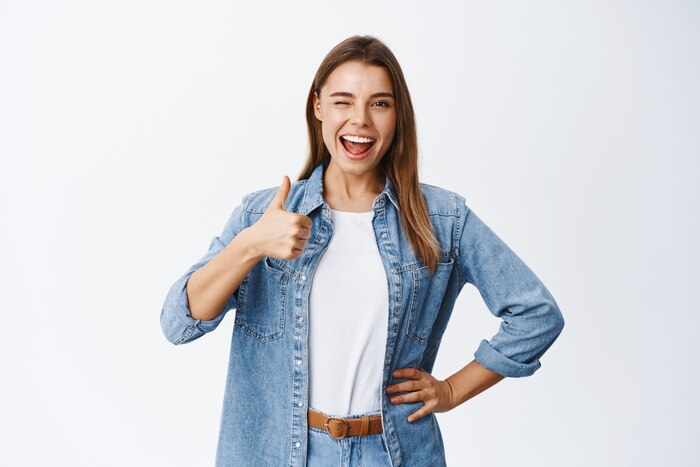 Cheerful young woman with blond hair and casual clothes, showing thumb up in approval, like good thing, approve and recommend it, praise nice job or saying yes, white wall