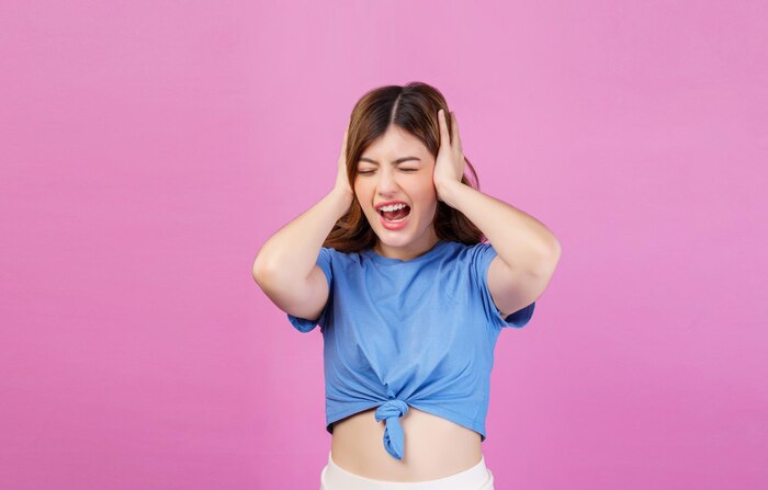 Portrait of angry irritated young woman wearing casual tshirt covering her ears with hands and shouting while standing isolated over pink background
