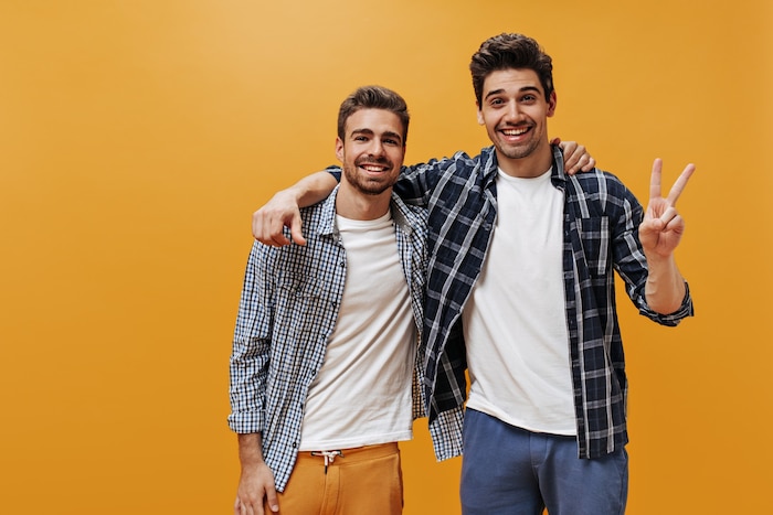 Cheerful young men in plaid blue shirts, white t-shirts and colorful pants pose on orange wall in great mood and smile.