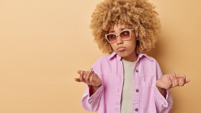 Hesitant young woman with curly hair shrugs shoulders in bewilderment feels doubt feels uncertain wears sunglasses and pink jacket isolated over beige background with empty space for promotion