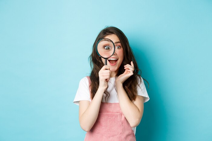 Happy young woman looking through magnifying glass with excited face, found or search something, standing over blue background
