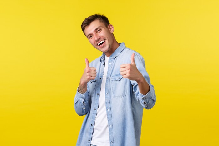 Portrait of handsome outgoing blond man showing thumbs-up in approval, encourage visit store. male student inviting people to summer event or courses with special discount, yellow background.