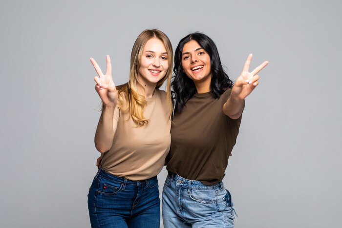 Portrait of a two girlfriends showing two fingers sign on gray wall and looking at front