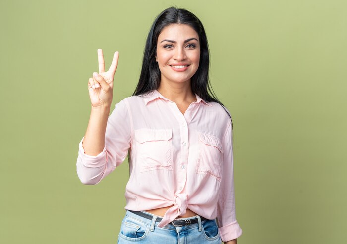 Young beautiful woman in casual clothes looking at front smiling cheerfully showing number two with fingers standing over green wall