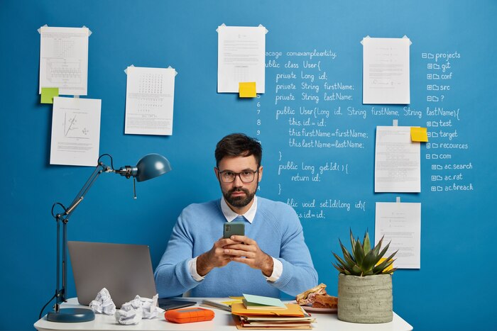 Digital technology expert or enthusiast obsessed with his work, uses mobile phone, works with modern devices, surrounded by many papers, poses at desktop