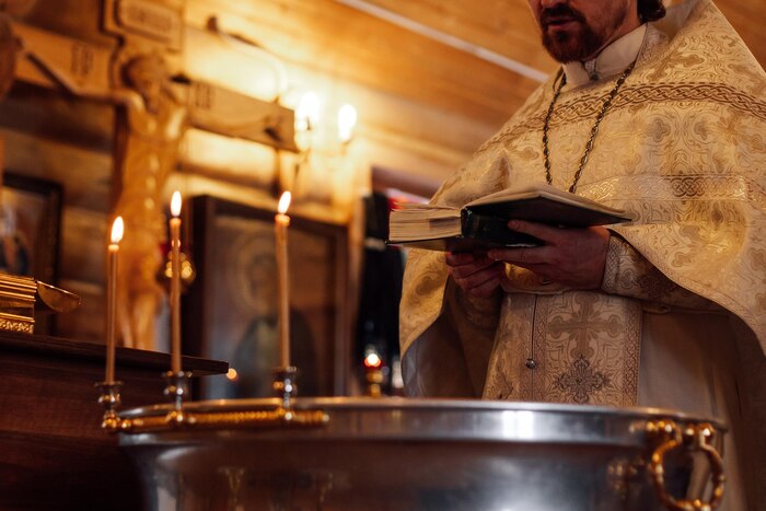 Cropped photo of priest wear ceremonial cassock standing at font decorated with burning candles reading prayer book
