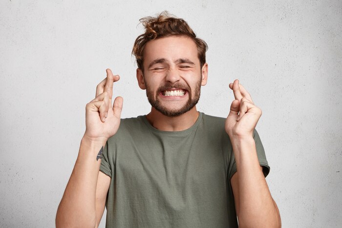 Cheerful bearded man keeps fingers crossed, smiles broadly and closes eyes