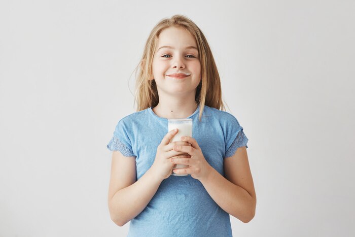Little cute girl with blonde hair in blue t-shirt with milk drops on face, happy to start her day with big glass of healthy drink.
