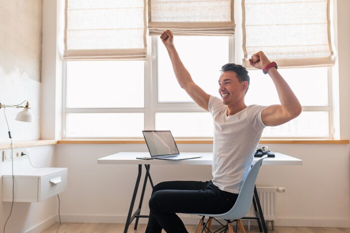 Young handsome smiling man in casual outfit sitting at table working on laptop, freelancer at home